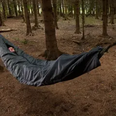 Mężczyzna śpi w czarnym śpiworze zawieszonym między drzewami w lesie. Spanie w hamaku to przygoda.