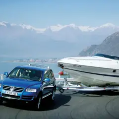 Un coche azul remolca un barco deportivo blanco por una carretera junto a un lago y montañas nevadas.