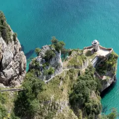 El faro del caballo se alza sobre un acantilado rocoso. Escaleras serpentean por la ladera verde hacia el mar turquesa.
