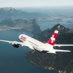 Samolot linii Helvetic Airways z charakterystycznym czerwonym ogonem i białym krzyżem leci nad malowniczym jeziorem i górami.