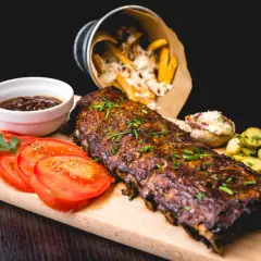 Un plat de côtes de porc au barbecue, garni de ciboulette, servi avec des tomates tranchées, des cornichons, une sauce barbecue et des frites au fromage.