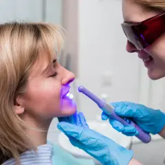 Dentystka w okularach ochronnych naświetla lampą zęby pacjentki. Dowiadujesz się, ile trwa wybielanie zębów.