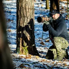 Fotografia przyrody: mężczyzna w kamuflażu kuca przy drzewie, celując profesjonalnym aparatem.
