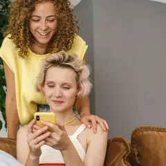 Dwie kobiety śmieją się, przeglądając telefon. Jedna z nich kładzie dłoń na ramieniu drugiej, sugerując wspólne konto dla par.