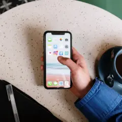 Main d'une personne tenant un smartphone affichant des icônes d'applications, comme un exemple d'application web. Une tasse de café est à côté.