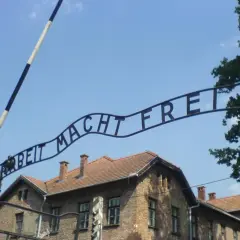 Brama z napisem "Arbeit Macht Frei" nad ceglanymi budynkami pod błękitnym niebem.