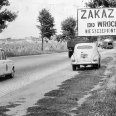 Znak "Zakaz wjazdu do Wrocławia osobom nieszczepionym przeciw ospie" przy drodze. W tle samochody i pola.