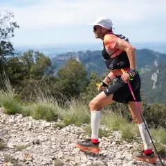 Uomo con bastoncini da trail running cos'è, corre su sentiero roccioso con vista panoramica su montagne e cielo azzurro.