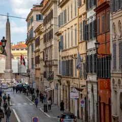 Una strada romana con palazzi storici, un obelisco e gente che passeggia. Si intuisce quanto è lunga Via del Corso, con la sua prospettiva che si perde all'orizzonte.