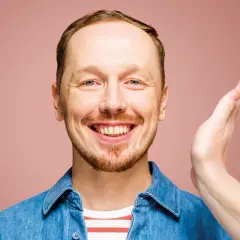 Uśmiechnięty mężczyzna w dżinsowej koszuli, jakby właśnie usłyszał świetny żart na występie syrek dąbrowski stand up.