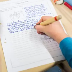 Kind schreibt auf liniertes Blatt. Kleine Zeichnungen illustrieren die Geschichte. Ein typischer Lernmoment im **Wortstamm Grundschule**.