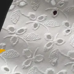 Close-up of white eyelet fabric with floral embroidery. This delicate fabric features cut-out holes surrounded by decorative stitching, creating a beautiful, airy pattern.