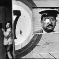 A man with a monocle and mustache, reminiscent of Cartier Bresson paintings, peers from behind a wooden structure. A blurred figure stands in the background.