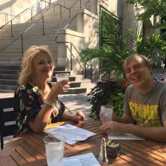 Tom Logan, mąż Majki Jeżowskiej, uśmiecha się do zdjęcia, siedząc przy stoliku na zewnątrz z drinkiem.