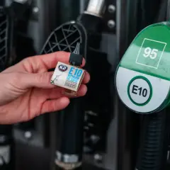 Dłoń trzyma małą buteleczkę z dodatkiem do paliwa E10 na tle dystrybutora paliwa z oznaczeniem 95 i E10.