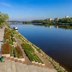 Nowoczesny boulevard nad rzeką z zielenią, nowoczesnym budynkiem i mostem w oddali.