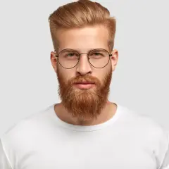Un homme au visage rectangulaire, barbe rousse et lunettes rondes, porte un t-shirt blanc.