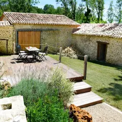 Aménagement longère : terrasse en bois avec mobilier, jardin verdoyant et murs en pierre.