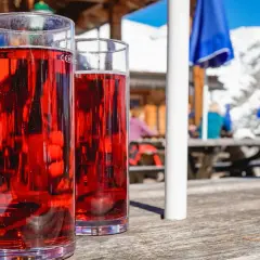 Zwei Gläser mit rotem Getränk, vielleicht ein Skiwasser-Rezept, stehen auf einem Holztisch. Im Hintergrund eine Skihütte mit Gästen.