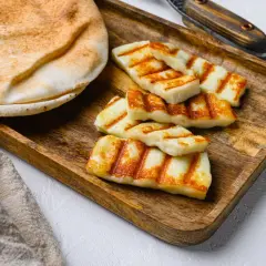 Des morceaux de halloumi grillé, marqués par les rayures du grill, reposent sur un plateau en bois à côté d'un pain pita moelleux.