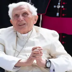 Emeritierter Papst Benedikt XVI. in weißer Soutane, Hände gefaltet, blickt lächelnd nach oben.
