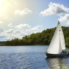Ahoj Mazury! Biały żagiel łodzi odbija się w spokojnej tafli jeziora, otoczonej zielonymi lasami. Słońce przebija się przez chmury.