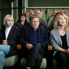 Małgorzata Englert, w wieku, siedzi w pierwszym rzędzie obok innych osób, wpatrując się w dal.