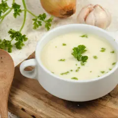 Aksamitna zupa krem z białych warzyw, udekorowana natką pietruszki, obok drewniana łyżka, cebula i czosnek.