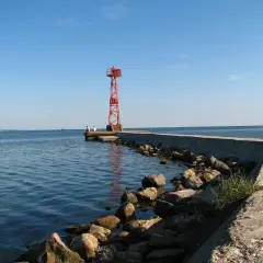 Czerwona wieża nawigacyjna na końcu falochronu w Jastarni. Dwie osoby siedzą na końcu molo, patrząc na spokojne morze.