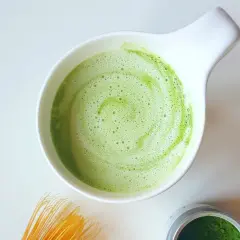 Zielony, spieniony napój matcha w białej miseczce, idealny jako alternatywa co zamiast kawy. Obok bambusowa miotełka i proszek matcha.