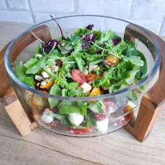 Pyszna sałatka z mix sałat, pomidorkami koktajlowymi, mozzarellą i orzeszkami pinii w szklanej misie na drewnianej podstawce.
