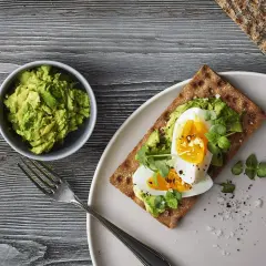 Delicioso sandwich de aguacate y huevo sobre pan integral, con un tazón de aguacate machacado y un tenedor al lado.
