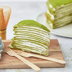 Ein Stück saftiger Matcha Kuchen mit vielen dünnen Schichten und cremiger Füllung, bestäubt mit grünem Pulver. Daneben ein Chasen und Holzgabeln.