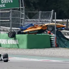 Wypadek bolidu F1 na torze Monza. Pomarańczowo-niebieski bolid leży na bandzie, ratownicy udzielają pomocy.