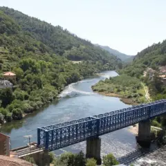 Un puente azul cruza un río serpenteante rodeado de colinas verdes y casas. El paisaje evoca la tranquilidad de los peares en un día soleado.