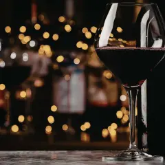 Szklanka z czerwonym winem, obok butelka. W tle migoczące światła, jakby wieczór w winiarni. To wino bycza moc.
