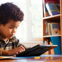 Ein Junge mit lockigem Haar lernt mit Bildungs-Apps auf einem Tablet. Im Hintergrund ein Bücherregal voller Bücher.