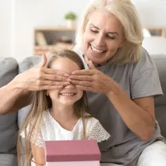 Babcia zasłania oczy wnuczce, która trzyma prezent. Radosna niespodzianka z okazji imienin Nikodema.