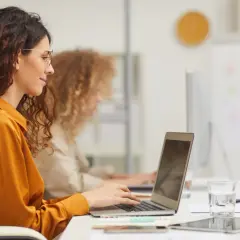 Kobieta w okularach pracuje przy laptopie, przygotowując umowę zlecenia. Obok szklanka wody i tablet.