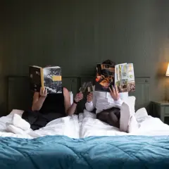 Två personer läser tidningar och skålar med champagne i en säng på Hotell Hertig Karl Filipstad.