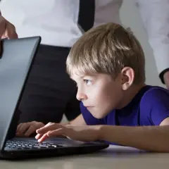 Chłopiec pochylony nad laptopem, nad nim ręka dorosłego.
