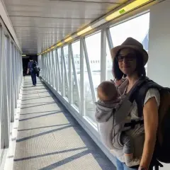 Kobieta z dzieckiem w chuście na rękawie lotniskowym, w tle samolot.