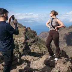 Un hombre fotografía el espectacular **barranco de Masca** mientras una mujer posa con las montañas y el mar de fondo.
