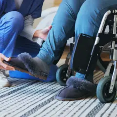 Opiekunka zakłada kapcie osobie na wózku. To codzienne świadczenie wspierające, pełne troski i empatii.