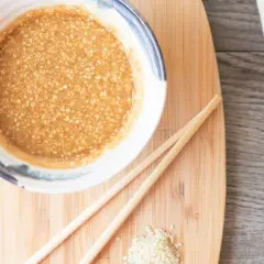 A bowl of creamy sesame sauce, perfect for your next sesame sauce recipe, sits on a wooden board with chopsticks and a sprinkle of sesame seeds.