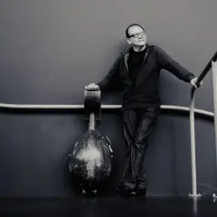 Anouar Brahem, w okularach, opiera się o poręcz, obok niego stoi instrument.