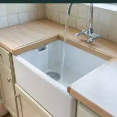 Biała, ceramiczna umywalka do kuchni z chromowaną baterią, w której płynie woda. Blat drewniany, szafki jasne.