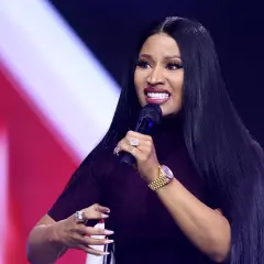 Nicki Minaj z mikrofonem, w ciemnej sukience, z długimi czarnymi włosami i biżuterią.