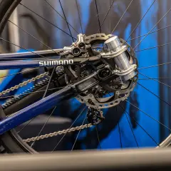 Close-up of a blue bicycle's rear wheel and Shimano drivetrain. A silver grip damper is visible on the axle, ready to absorb shocks.