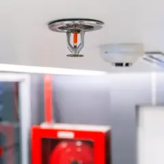 Eine Sprinkleranlage an der Decke, bereit für den Notfall. Daneben ein Rauchmelder und ein grünes Notausgangsschild.